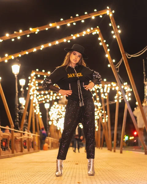 街のクリスマスライト 黒とスパンコールの服を着た白人ブロンドの女の子 ライトアップされた橋の上の帽子 冬のライフスタイル — ストック写真