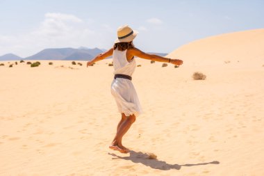 Beyaz elbiseli ve şapkalı genç bir Kafkasyalı turist, Kanarya Adaları, Fuerteventura 'daki Corralejo Doğal Parkı' nın kumullarında yürüyor. İspanya