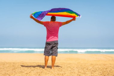 Pembe tişörtlü, siyah şapkalı, kumsalda LGBT bayrağı olan eşcinsel biri. Homoseksüelliğin sembolü.