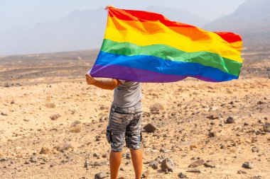 Gri tişörtlü, siyah şapkalı, LGBT bayrağı rüzgarlı bir çölde sallanan eşcinsel bir adam. Homoseksüelliğin sembolü.