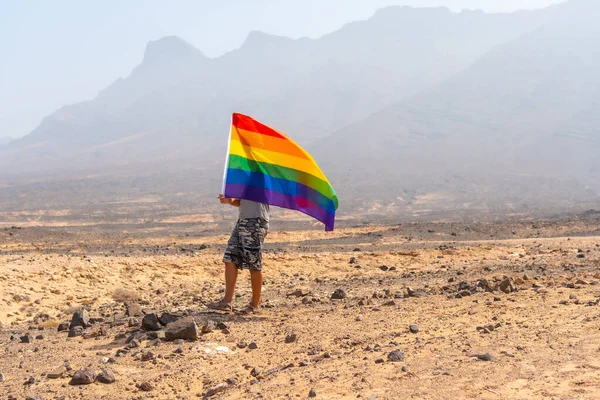 Gri tişörtlü, siyah şapkalı, LGBT bayrağı rüzgarlı bir çölde sallanan, eşcinselliğin sembolü olan tanınmamış bir gay.