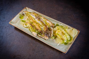 Yeşil soslu balık, siyah arka planda patates, beyaz tabakta.