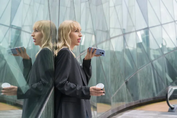 現代的な建物の中にある起業家の女の子のビジネスパークは 窓に反映され 若いブロンドのスーツは テイクアウトコーヒーと音声メッセージを送信 — ストック写真
