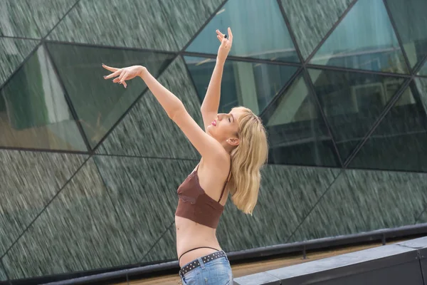 Cam bir binada poz veren genç sarışın, kahverengi tişörtlü ve kot pantolonlu sarışın kız, kollarını kaldır.