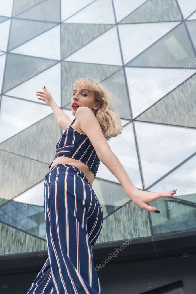 Fashionable posing of a young blonde in a glass building, blonde girl in a blue striped suit, arms up, vertical photo