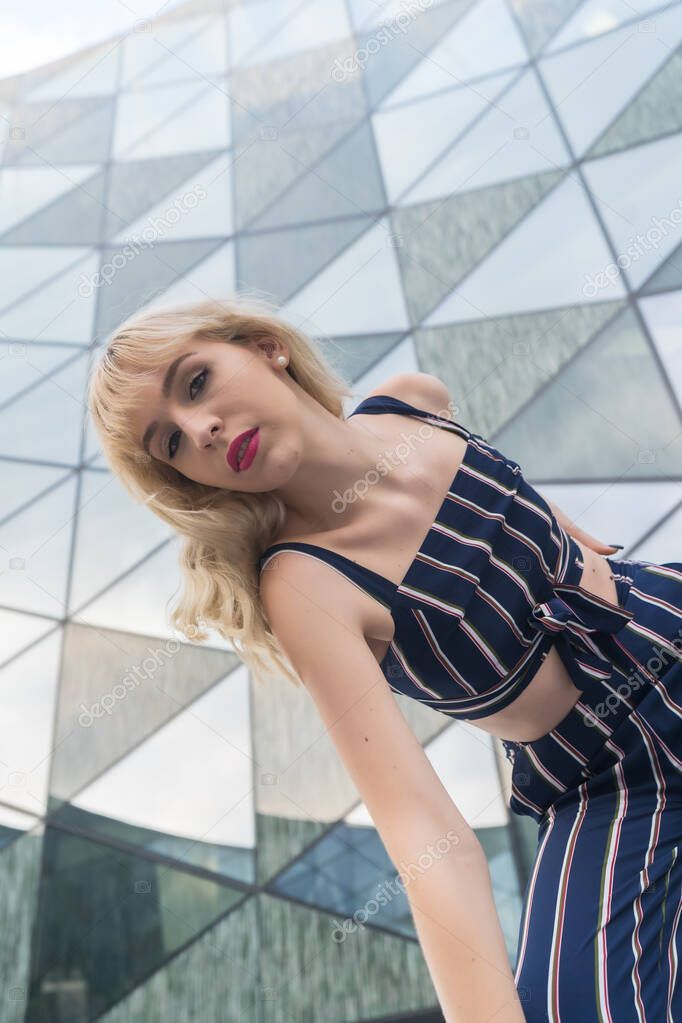 Below photo of a young model in a glass building, blonde girl in a blue striped suit, looking towards the camera, vertical photo