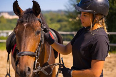Beyaz, sarışın bir kız, at üstünde kahverengi bir atı okşuyor ve şımartıyor. Siyah atlı, güvenlik şapkası takıyor.