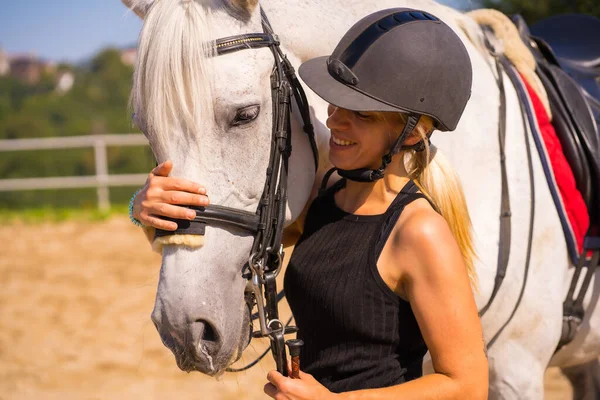 白人ブロンドの女の子馬に乗って白い馬の頭を抱いて 安全帽をかぶった黒いライダーに身を包んだ — ストック写真
