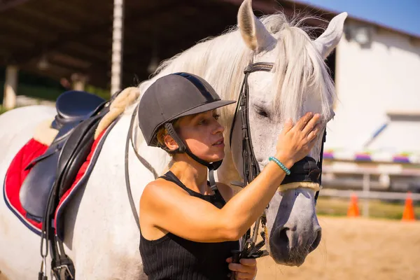 白人ブロンドの女の子は安全帽をかぶった黒のライダーで身を包んだ乗馬で白い馬を撫でて — ストック写真