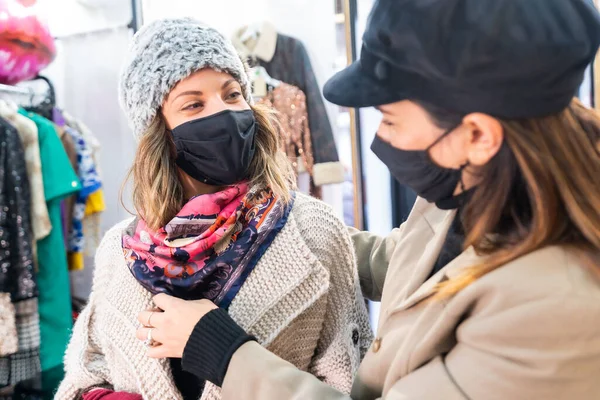 Caucasian employee with a mask from a clothing store testing a customer a pink scarf in a mirror, security measures in the coronavirus pandemic, covid-19