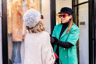 Şehirde alışveriş yapan genç beyaz bir kız ve giyim mağazalarının vitrinine bakan bir tezgahtar tarafından bakılıyor. Güneş gözlüğü ve kırmızı eldivenler