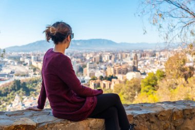 A young tourist sitting looking at the views of the city and the Cathedral of the Incarnation of Malaga from the Castillo de Gibralfaro in the city of Malaga, Andalusia. Spain