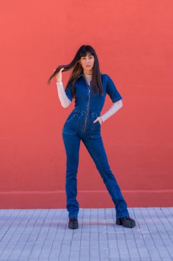Mavi kot elbiseli, duvara yaslanmış gülümseyen genç, güzel bir esmer kız. Kırmızı arka planda güzel bir beyaz poz
