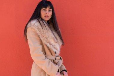 Elinde bir çantayla duvara yaslanmış kışlık ceketli, gülümseyen esmer bir manken pozu. Beyaz, güzel, kırmızı bir arka plan.