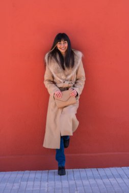 Kışlık ceketli gülümseyen esmer kız bir çantayla duvara yaslanıyor. Beyaz, güzel, kırmızı bir arka plan.