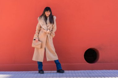 Elinde bir çantayla duvara yaslanmış kışlık ceketli, gülümseyen esmer bir manken pozu. Beyaz, güzel, kırmızı bir arka plan.