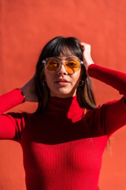 Elleri yüzlerinde turuncu bir elbise ve güneş gözlüğü takan esmer bir kızın portresi güneşli bir sabahın tadını çıkarıyor. Beyaz, güzel, kırmızı bir arka plan.