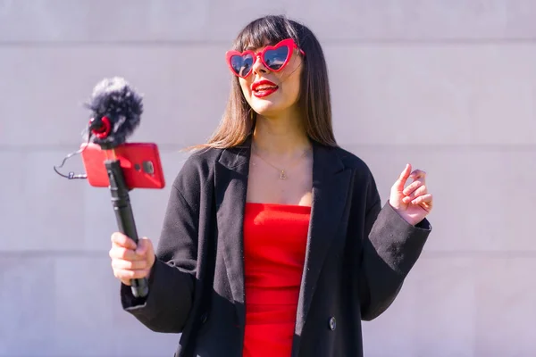 ブルネットCaucasian女の子作るA Vlog Valentineの日のためのソーシャルネットワーク 赤いハートサングラス 電話とマイクで話す灰色の背景 — ストック写真