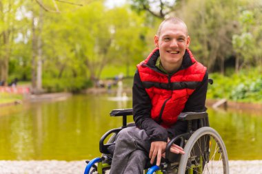 Şehir parkında felçli bir gencin portresi. Tekerlekli sandalyede oturmuş kameraya bakarak gülümsüyorum.