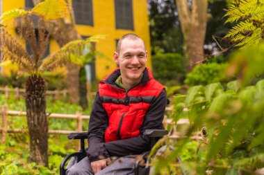 Şehir parkında kırmızı yelek giyen felçli bir gencin portresi. Tekerlekli sandalyede oturmuş gülümsüyor.