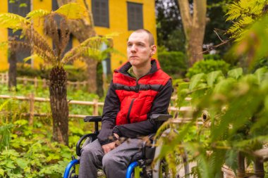 Şehir parkında kırmızı yelek giyen felçli bir gencin portresi. Tekerlekli sandalyede oturuyor.