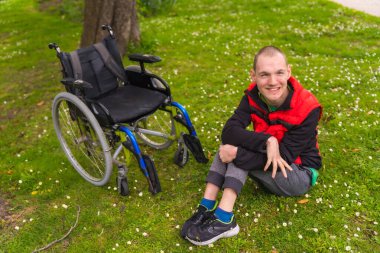 Doğal bir parkta, baharda tekerlekli sandalyenin yanındaki çimlerde oturan felçli bir gencin portresi.