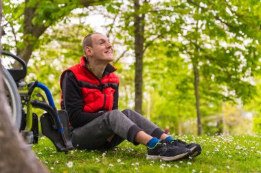 Kötürüm bir gencin portresi. Çimlerin üzerinde, tekerlekli sandalyenin yanında. Bir ağacın yanında.