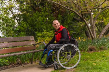 Şehir parkında kırmızı yelek giyen felçli bir gencin portresi. Tekerlekli sandalyede bir park sandalyesinin yanında oturuyor.