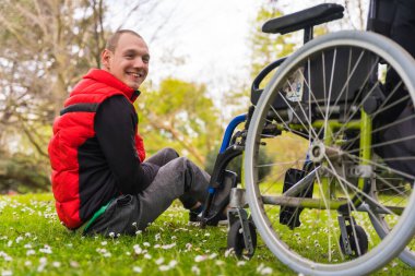 Felç olmuş genç bir adam, tekerlekli sandalyenin yanındaki çimenlerde oturuyor, bir ağacın yanında kameraya bakıyor ve gülümsüyor.