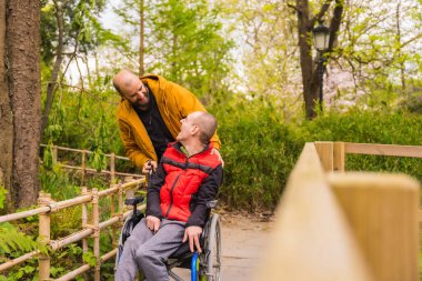 Tekerlekli sandalyedeki felçli genç adam halk parkında birbirine bakıp gülümseyen bir arkadaş tarafından itiliyor.