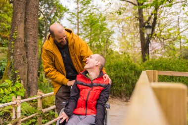 Tekerlekli sandalyedeki felçli genç adam halk parkında birbirine bakıp gülümseyen bir arkadaş tarafından itiliyor.