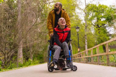 Tekerlekli sandalyedeki felçli genç bir adam halk parkında bir arkadaş tarafından itiliyor, bir yolda yürüyor, eğleniyor ve gülümsüyor.