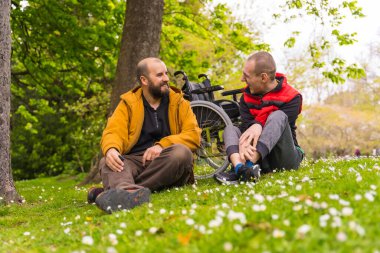 Felç olmuş genç bir adam ve bir arkadaşı şehirdeki bir halk parkının çimlerinde oturuyor, konuşuyor ve gülüyorlar.