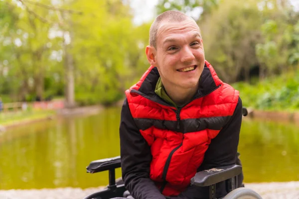 Şehir parkında felçli bir gencin portresi. Tekerlekli sandalyede oturmuş kameraya bakarak gülümsüyorum.
