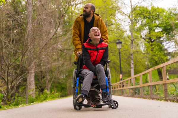 Tekerlekli sandalyedeki felçli genç bir adam halk parkında bir arkadaş tarafından itiliyor, bir yolda yürüyor, eğleniyor ve gülümsüyor.