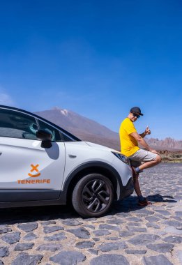 Genç bir adam Tenerife, Kanarya Adaları 'ndaki Teide Doğal Parkı' ndaki Zapato de La Reina perspektifinde kiralık arabanın yanında poz veriyor.
