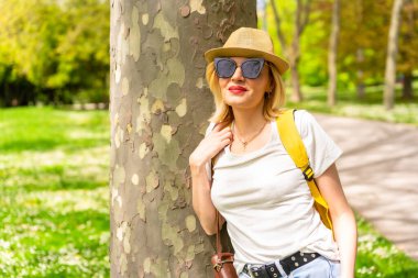 Sarışın, şapkalı ve güneş gözlüklü bir turist. İlkbaharda şehrin parkında bir ağaca yaslanmış, elinde kamerayla güneşin tadını çıkarıyor.