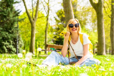 Sarışın turist bir kadın telefonuyla ses notu yolluyor, şapka takıyor ve güneş gözlüğü takıyor baharda çimlerin üzerinde şehrin parkında papatyaların yanında oturuyor.
