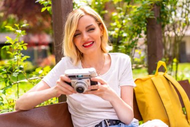 Şehirdeki bir parkta tatilde çektirdiği fotoğrafların keyfini çıkaran turist bir kız, bankta oturan bir şapka takıyor.