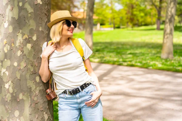 今年春天 在城市的一个公园里 一位戴着帽子和太阳镜的金发女游客靠着一棵树 带着相机 向右看去 — 图库照片