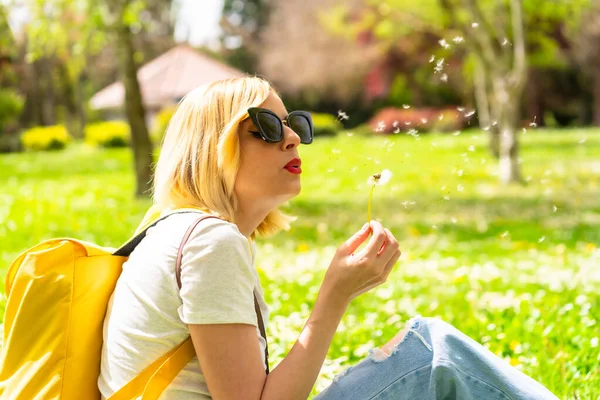 Turystka Blondynka Dmuchająca Mniszek Lekarski Kapeluszu Okularach Przeciwsłonecznych Siedząca Trawie — Zdjęcie stockowe