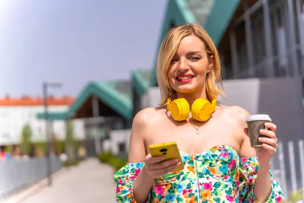 Portret Blondynki Chodzącej Telefonem Przez Miasto Żółtymi Słuchawkami Kawą Wynos — Zdjęcie stockowe