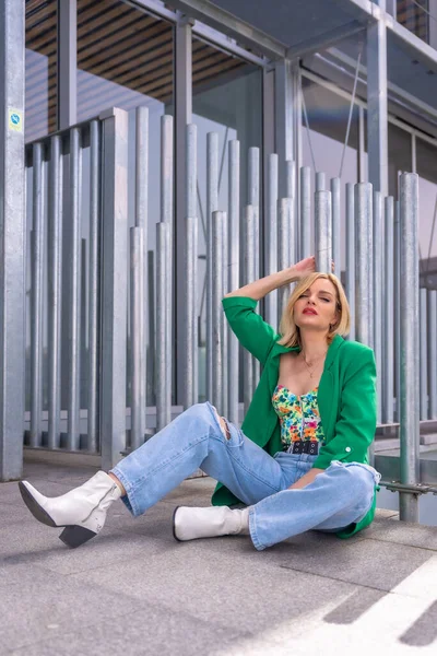 Şehirde bir binanın zemininde oturan yeşil ceketli sarışın model bir kadının portresi. Yaz tatili yaşam tarzı