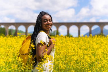 Tatilin tadını çıkaran, sarı çiçeklerle dolu bir tarlada örgülü, gezgin siyah etnik kız.