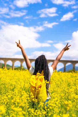 Zafer sembolünü yapmak için, sarı çiçekli bir tarlada örgülü, gezgin siyah etnik bir kız.