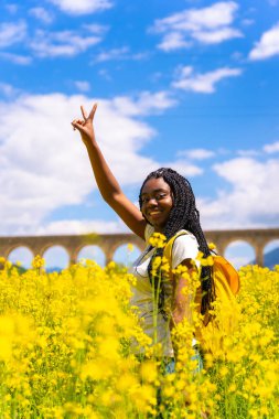 Zafer işareti yapan, örgülü siyah etnik bir kız, bir gezgin, sarı çiçeklerle dolu bir tarlada