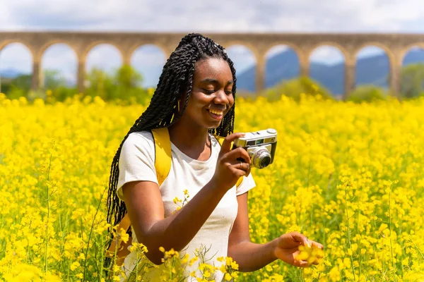 Eski model bir kamerayla çiçeklerin fotoğraflarını çekmek, sarı çiçekli bir tarlada örgülü siyah bir etnik kız, bir gezgin,