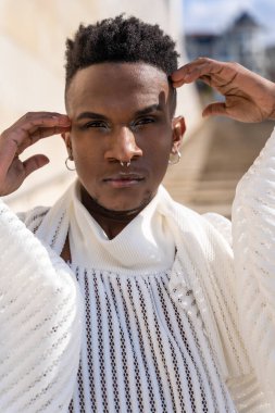 Beyaz takım elbiseli, siyah deri botlu, Afrika kökenli genç bir adam, şehirde şık bir model tuğla bir arka planda, yüzünde ellerle portresi, baştan çıkarıcı bir görünüm.