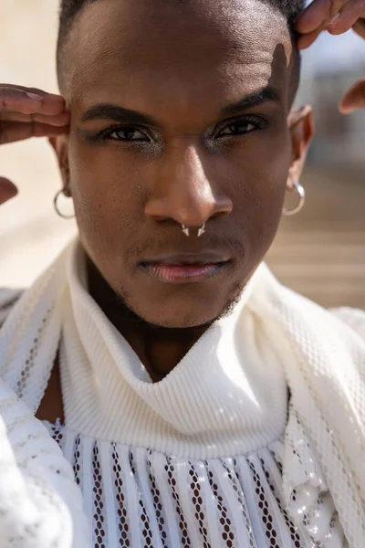 Beyaz takım elbiseli, siyah deri botlu, Afrika kökenli genç bir adam, şehirde şık bir model tuğla bir arka planda, yüzünde ellerle portresi, baştan çıkarıcı bir görünüm.