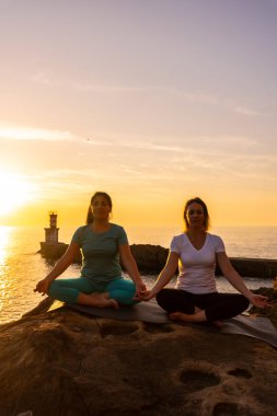 İki kadın gün batımında deniz kenarında yoga minderinde oturuyor, sağlıklı ve natürist yaşam tarzı, açık hava pilatesi, ardha padmasana